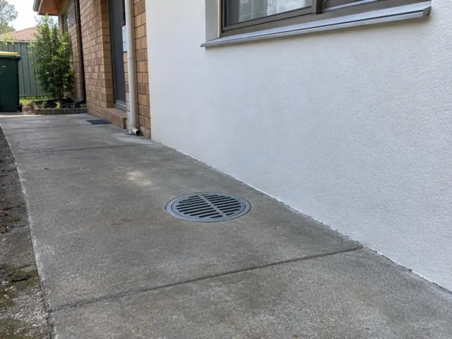 Round outdoor drain grate set in a concrete side path beside a house wall in Canley Heights