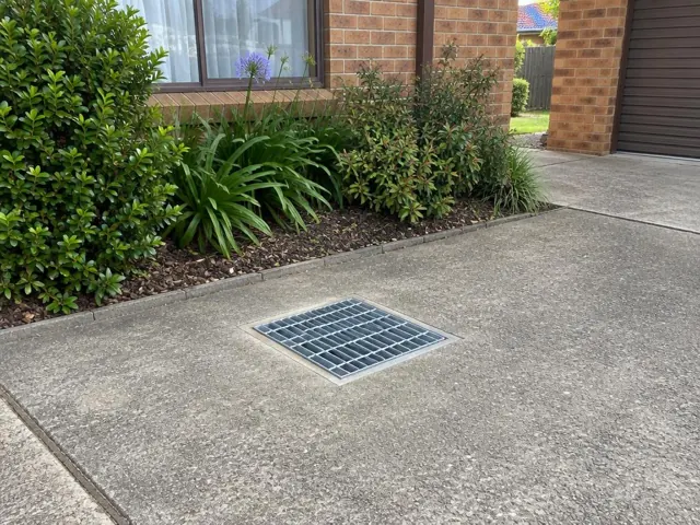 Flush driveway stormwater grate beside brick house and garden in Carramar